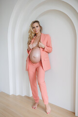 Beautiful pregnant woman in orange peach color suit, trousers and blazer. Young blond lady in spacious room with lamps and window. Big pregnant naked belly. Pretty woman waiting child.