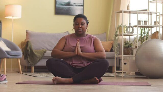 Stab shot of young curvy African American woman in activewear sitting on yoga mat in bright living room meditating