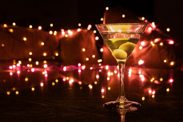 A clear glass of martini with olives focuses on a olives on a night club setting, and blurred background light