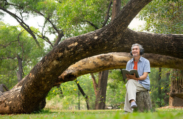 Naklejka na ściany i meble senior man with grey hair holding a book, wearing headphone to listen, smiling, sitting near a big tree in forest park. concept elderly health benefits in nature,wellbeing