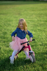 Fototapeta premium A happy beautiful little girl runs fast and rides her first bicycle without pedals on the grass on a warm summer day. Cute baby 3 years old. Side view. Learning to keep balance.