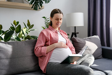 Happy young caucasian woman in advanced pregnancy relaxes on maternity leave sitting on comfy couch, spend free time reading book, gently strokes belly, feeling baby kicking. Motherhood concept