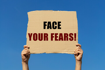 Face your fears text on box paper held by 2 hands with isolated blue sky background. This message board can be used as business concept about facing your fears.