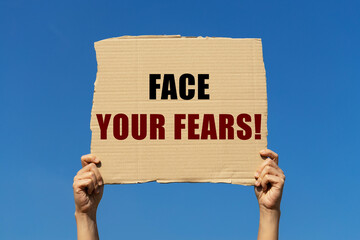 Face your fears text on box paper held by 2 hands with isolated blue sky background. This message...