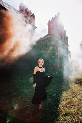 Art work. A slender beautiful woman with blond hair in a black vintage dress and expensive jewelry, stylish makeup and hairstyle, comes out of an ancient castle in fire and smoke.