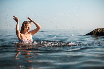 A blonde girl with long hair, in a striped swimsuit, bathes in the ocean, laughs, plays and splashes on a bright sunny day.