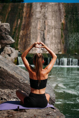 A tanned girl with an athletic figure sits in a lotus position and shows the shape of a heart with her fingers, against the backdrop of a picturesque dam. She is wearing black shorts and a top.