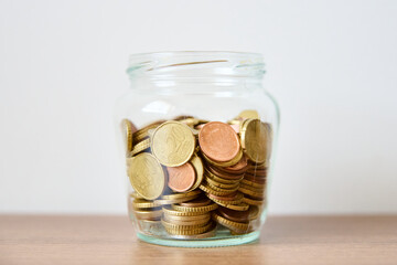Coins in glass jar on wooden table and white wall background.