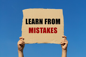 Learn from mistakes text on box paper held by 2 hands with isolated blue sky background. This message board can be used as business concept about lesson learned.