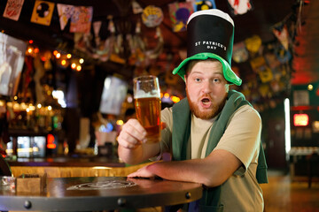 A smiling happy man in a leprechaun hat with beer in pub. He celebrates St. Patrick