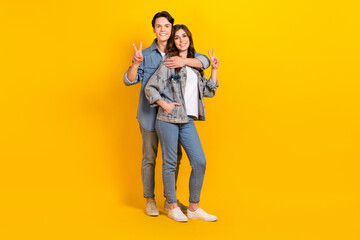 Full length photo of sweet pretty couple wear jeans showing v-signs cuddling isolated yellow color background