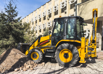 A small yellow excavator is digging a trench near a city building.