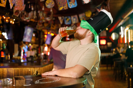 A man in a leprechaun hat and with a beard drinks beer in a bar