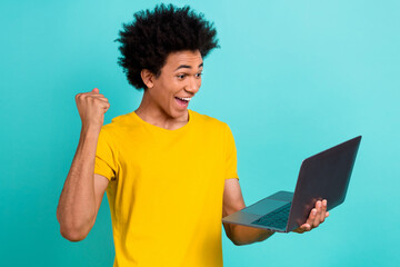 Portrait of ecstatic satisfied guy wear yellow t-shirt hold laptop raising fist celebrate win isolated on vibrant teal color background