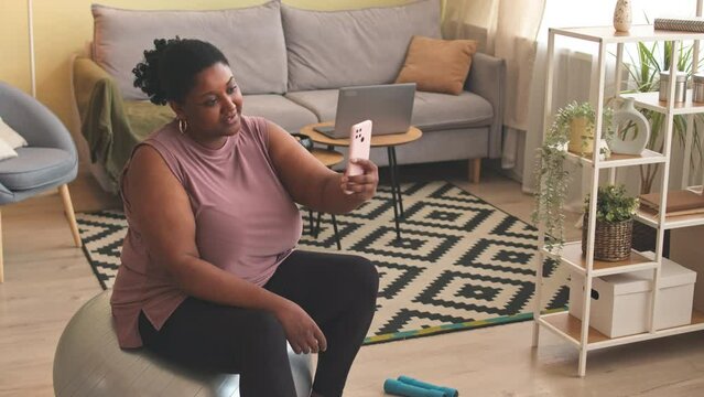 Young smiling curvy Black woman sitting on fitness ball and taking selfie portrait on smartphone while training at home