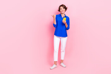 Full body photo of young positive influencer girl wear stylish clothes hold phone point finger empty space website isolated on pink color background © deagreez