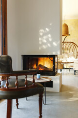 Cup of hot drink on table, warm fireplace with armchair in front and natural light from window