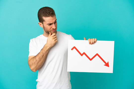 Handsome blonde man over isolated blue background holding a sign with a decreasing statistics arrow symbol and thinking