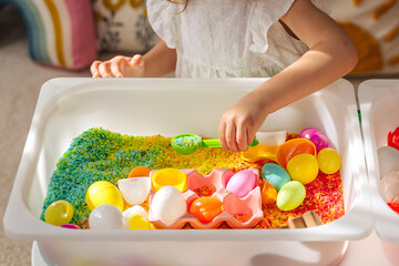 A little girl playing with colored rice and Easter eggs in sensory bin. Easter sensory bin with bright rice and eggs, bunny, carrot. Sensory play and holidays activity for kids. Happy Easter concept. - obrazy, fototapety, plakaty