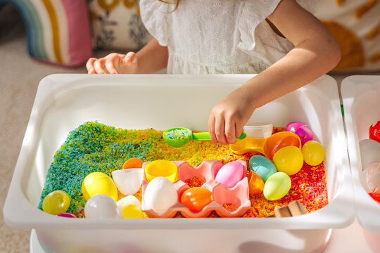 A little girl playing with colored rice and Easter eggs in sensory bin. Easter sensory bin with bright rice and eggs, bunny, carrot. Sensory play and holidays activity for kids. Happy Easter concept.