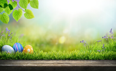 Three painted easter eggs celebrating a Happy Easter on a spring day with a green grass meadow, bright sunlight, tree leaves and a background with copy space and a wooden bench to display products. - obrazy, fototapety, plakaty