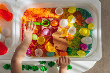 Child playing with colored rice and Easter eggs in sensory bin. Easter sensory bin with bright rice and eggs, bunny, carrot. Sensory play and holidays activity for kids. Happy Easter concept.