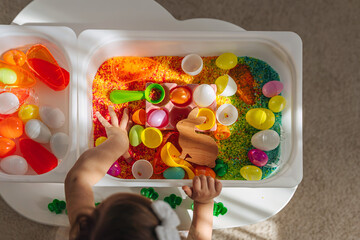 Child playing with colored rice and Easter eggs in sensory bin. Easter sensory bin with bright rice and eggs, bunny, carrot. Sensory play and holidays activity for kids. Happy Easter concept. - obrazy, fototapety, plakaty