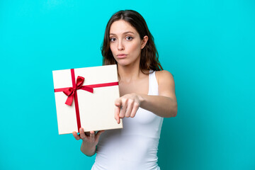 Young caucasian woman holding a gift isolated on blue background surprised and pointing front