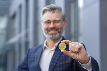 Close-up photo. Portrait of happy senior gray-haired man, businessman standing outside office and holding gold coin in hand. Presents and shows bitcoin to the camera.