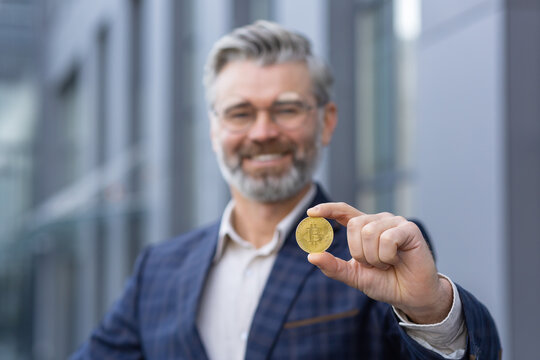 Close-up photo. Portrait of happy senior gray-haired man, businessman standing outside office and holding gold coin in hand. Presents and shows bitcoin to the camera.