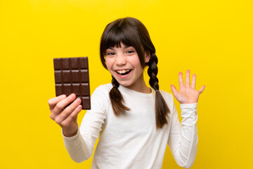 Little caucasian girl isolated on yellow background taking a chocolate tablet and surprised