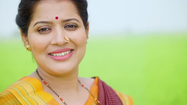 Close up shot of happy smiling traditional Indian woman looking at camera at green farmland - concept of individuality, elegance and satisfaction.