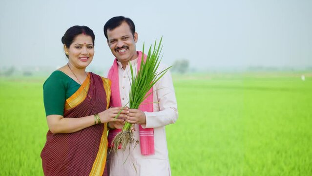 Happy smiling farming village couple standing by holding paddy crop while looking camera at farmland - concept of successful cultivation, harvesting season and bumper crop yield.