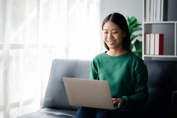 Naklejka premium A beautiful Asian woman sits on the sofa in the living room, she is using her laptop to play social media and chat with friends online. The concept of using technology to communicate on the Internet.