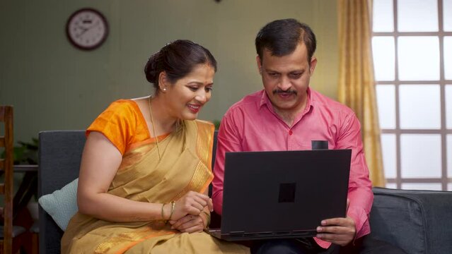 Happy middle aged couple discussing by watching laptop while sitting on sofa at home - concept of new home interior planning, checking apartments and online surfing.