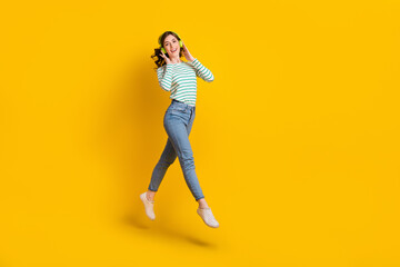 Full length photo of good mood nice girl wear striped shirt denim pants flying touching headphones isolated on yellow color background