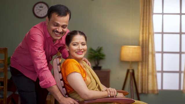Happy smiling middle aged couple looking at camera while sitting on chair at home - concept of successful relationship, family bonding and supportive husband.