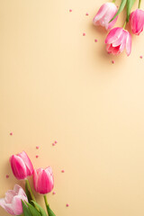Mother's Day celebration concept. Top view vertical photo of bunches of pink tulips and heart shaped sprinkles on isolated pastel beige background with copyspace