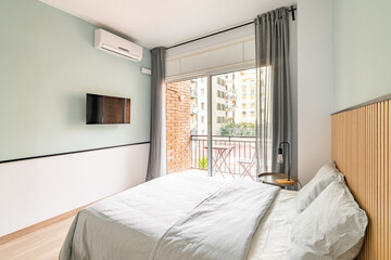 Small but pleasant bedroom with a double bed with white linens, TV and an open terrace overlooking the courtyard of a residential complex or tourist hotel on a sunny summer day