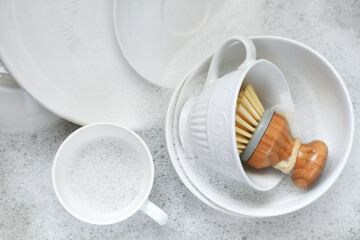 White dishes plates mugs in sink of dishwater with soap bubbles and scrub brush