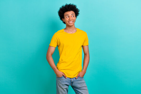 Portrait of good mood guy afro hairdo wear yellow t-shirt look empty space hold hands in pockets isolated on shine teal color background