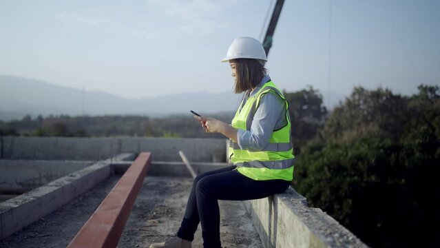 Civil engineer woman sitting and use the phone to contact work.on Industrial building. Architect on construction site. Construction and architecture. Professional engineer in safety equipment. Reflect