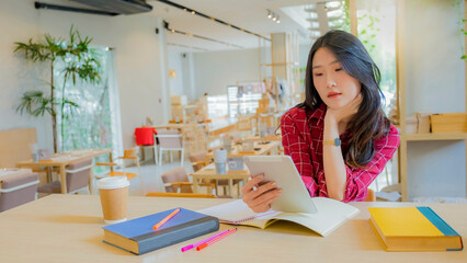 Young smiling asian woman with long brown hair, dressed in a red white striped shirt, is holding a tablet computer and a pen at a study desk. beautiful sitting at cafe she relaxed. out of focus.