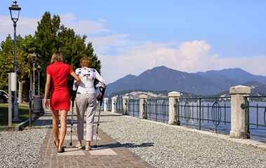 Young caregiver with elderly woman who uses crutches because she can't walk due to walking problems. Walk on the shore of Lake Maggiore in Italy.