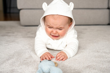 Authentic lifestyle photo of infant baby girl seven months old which lying on floor at home and crying because feeling sadness, hungry or pain