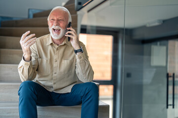 Gray haired senior man laughing while talking on smart phone, sitting on staircase in office lobby near modern glass office. Satisfied senior on entertaining cell phone conversation looking happy.