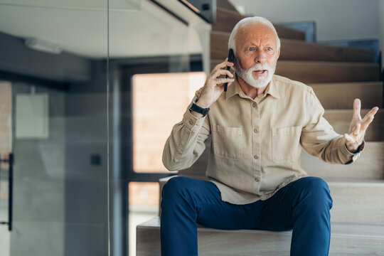 Senior Man Talking On Mobile Phone, Looking Upset And Extremely Nervous About Hearing The Bad News Over Phone Call, Sitting Indoors On Stairs, Gesturing With Hand And Looking Away.