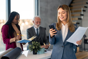 Beautiful stylish businesswoman in suit holding documents using mobile phone technology checking...