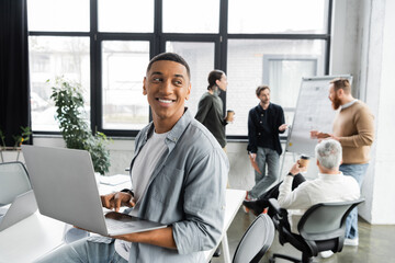 Cheerful african american businessman using laptop near blurred colleagues working in office. - obrazy, fototapety, plakaty