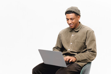 Young handsome african-american freelancer guy working with laptop isolated on white. Hipster man in shirt and a hat is holding a laptop while sitting in a chair and tping